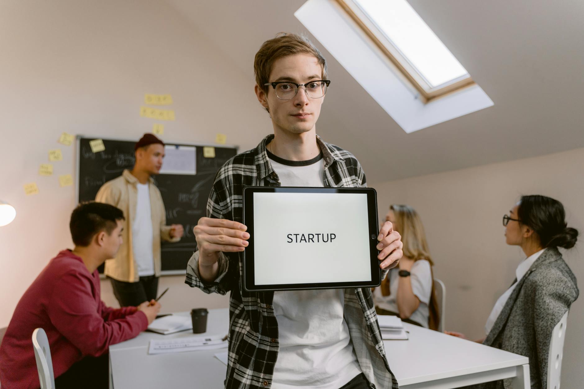 A young man in an office holding a tablet with 'STARTUP' during a meeting.