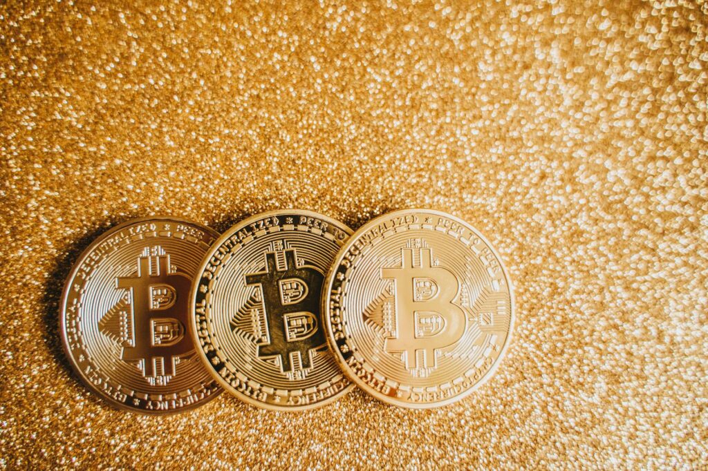 Close-up of three shiny Bitcoin coins on a sparkling gold glitter background, symbolizing cryptocurrency wealth.