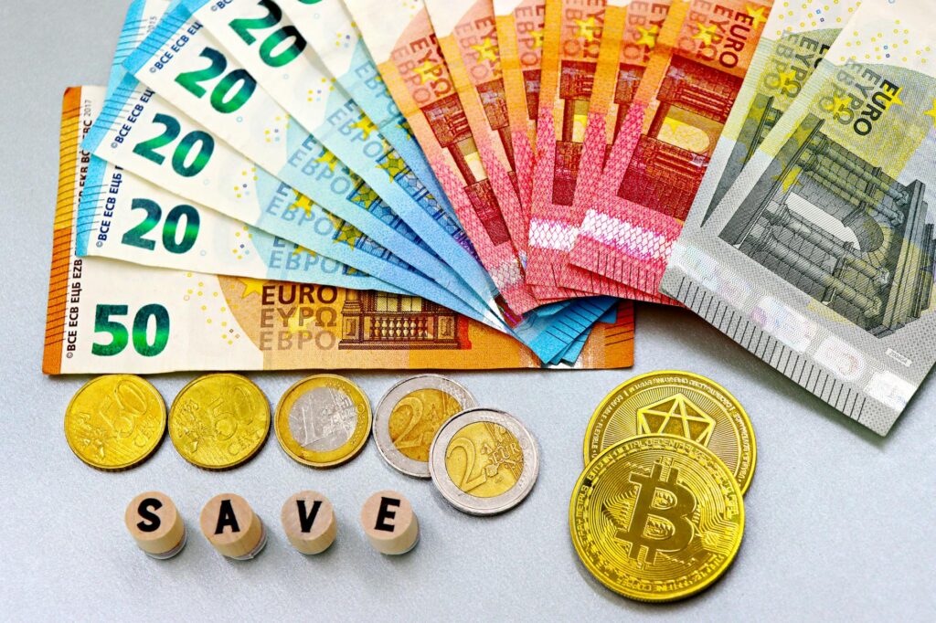 Euro banknotes and cryptocurrency coins arranged on a white surface with blocks spelling 'SAVE'.