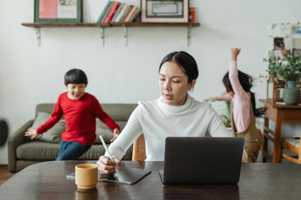 Asian mother working online using laptop and taking notes on tablet with stylus and cheerful children making noise and running behind in living room