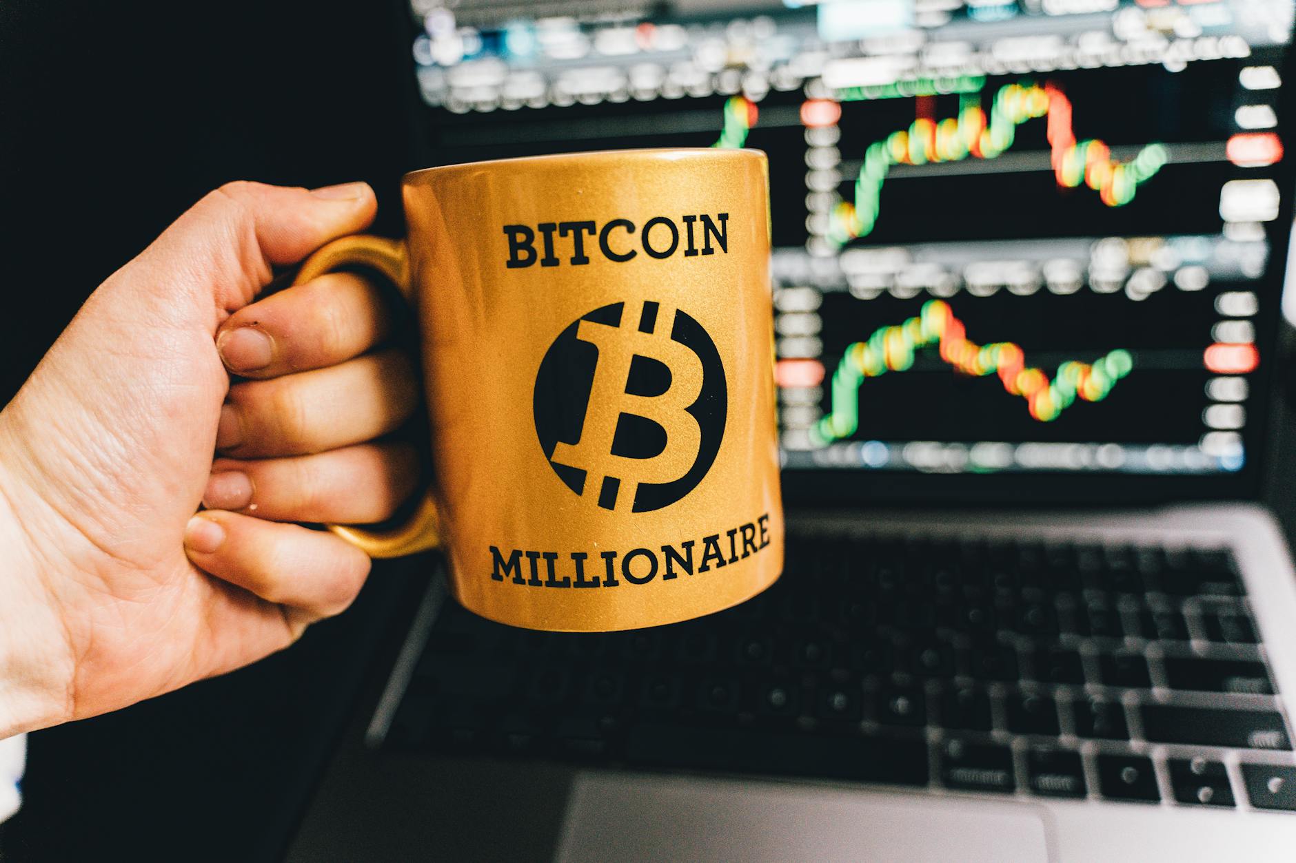 Golden mug with 'Bitcoin Millionaire' text in front of stock market charts on laptop screen.
