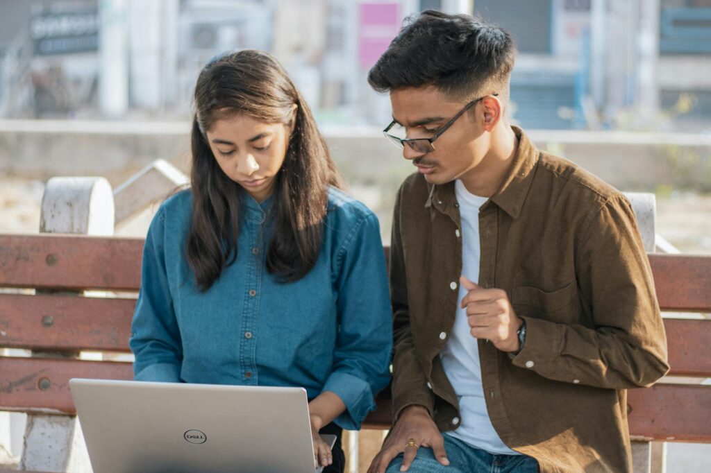 Two young adults working together on a laptop while sitting on a park bench outdoors.