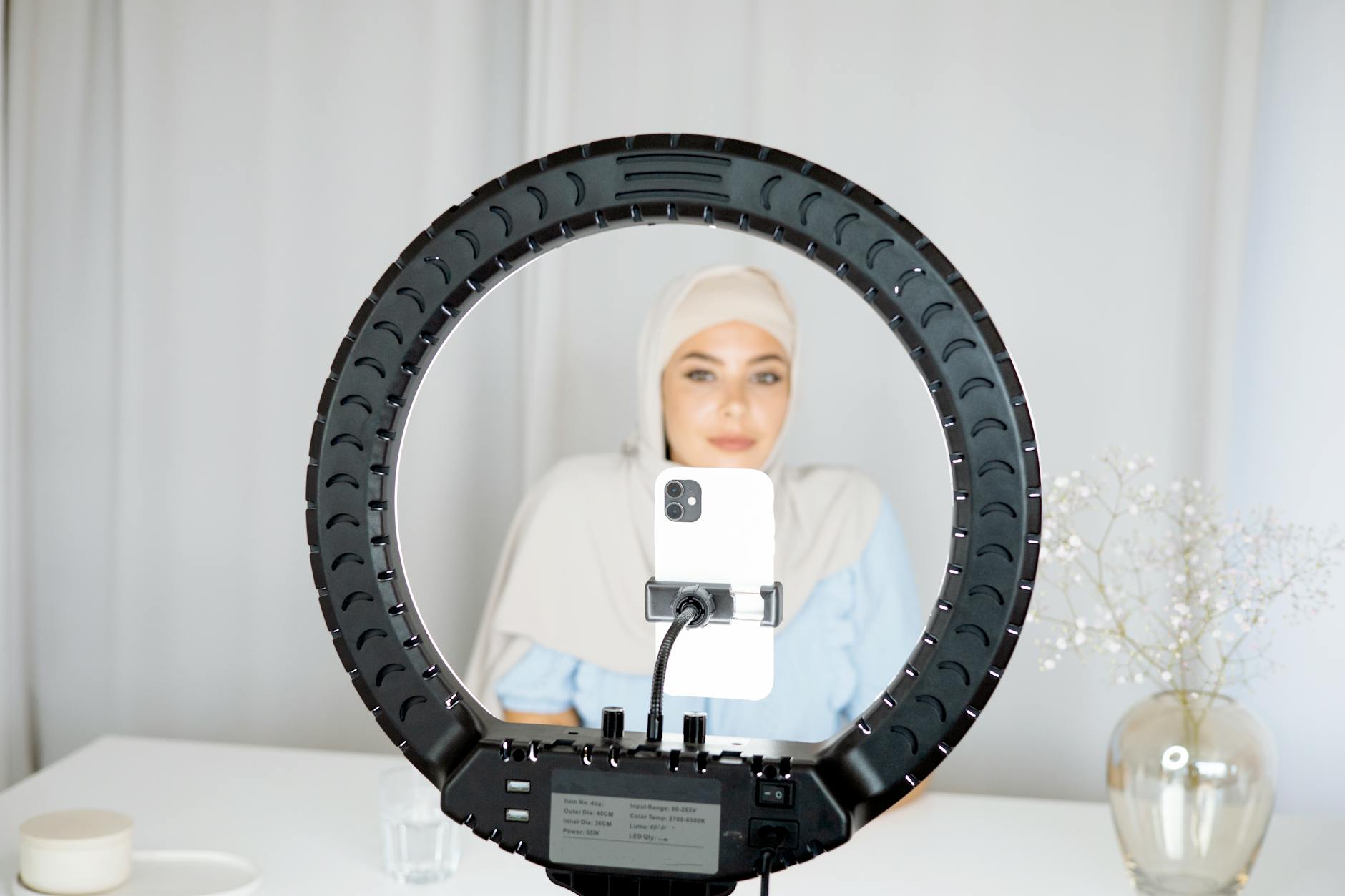 A woman in hijab using a ring light to create content indoors with a smartphone.